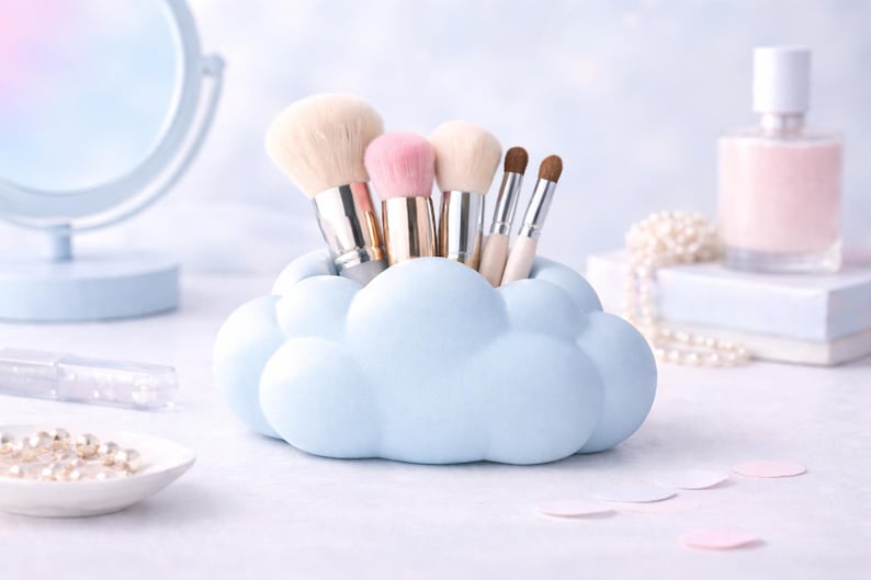 K&ouml;nnte beinhalten: Ein hellblaues, wolkenf&ouml;rmiges Make-up-Pinsel-Halter mit verschiedenen Make-up-Pinseln. Die Pinsel haben hellfarbene Borsten und metallische Griffe. Der Hintergrund umfasst einen Spiegel, Parf&uuml;m und Perlen, was eine weiche, pastellfarbene &Auml;sthetik erzeugt.