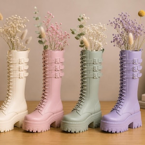 May include: Four tall, lace-up platform boots in cream, light pink, sage green, and lavender, each functioning as a vase. The boots feature three horizontal buckle straps at the top and thick, lugged soles, with dried flowers arranged inside each.