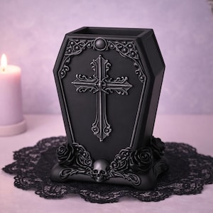 May include: Black coffin-shaped decorative container with a cross design. The container has ornate detailing, including roses and a skull at the base. It sits on a black lace doily, with a lit candle and a decorative box in the background.