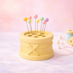 Peut inclure: Un coussin à épingles jaune clair avec un motif en croix, contenant des épingles à tête perlée colorées. Des bobines de fil rose et bleu, ainsi que des perles en vrac, sont disposées sur une surface rose et blanche, suggérant un thème de couture ou d'artisanat.