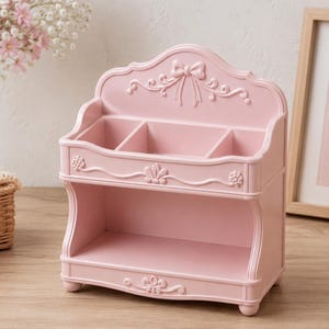 May include: A light pink decorative organizer with three compartments and a lower shelf. The organizer has ornate detailing, including a bow and floral accents. A framed picture and a vase of flowers are in the background.