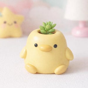 Puede incluir: Una maceta amarilla con forma de pollito con una pequeña suculenta verde. La maceta tiene ojos negros y un pico amarillo. Una figura en forma de estrella amarilla está en el fondo. Una lámpara rosa también está en el fondo.