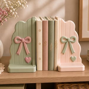 Puede incluir: Un par de sujetalibros arqueados con diseño de paneles verticales. El sujetalibros izquierdo es verde salvia con un lazo y un corazón rosa, mientras que el derecho es rosa pálido con un lazo y un corazón verde salvia. Sostienen cuatro libros.