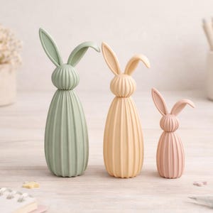 May include: Three pastel-colored bunny figurines. The bunnies are in shades of green, peach, and pink. Each has long ears and a ribbed body. They are arranged on a light-colored wooden surface, suggesting a spring or Easter theme.