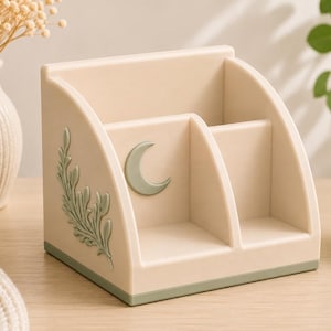 Op de afbeelding: Een beige bureau-organizer met drie vakken en een saliegroene basis. Het zijpaneel is voorzien van een verhoogd saliegroen bladmotief, terwijl het voorste middenpaneel een verhoogd saliegroen maansikkel-embleem op een glad oppervlak heeft.