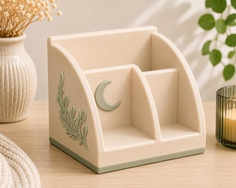May include: A beige desk organizer featuring three compartments with a sage green base. The side panel displays a raised sage green leaf motif, while the front center panel features a raised sage green crescent moon emblem on a smooth surface.