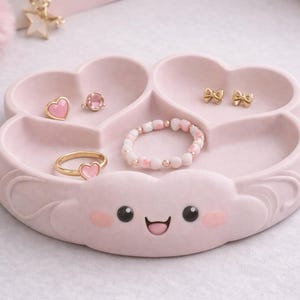 Può includere: Un vassoio portagioie rosa chiaro a forma di nuvola con scomparti a forma di cuore. Il vassoio contiene un anello d'oro con un cuore rosa, un orecchino a forma di cuore rosa, un orecchino a bottone con una gemma rosa, un braccialetto di perline e orecchini a fiocco dorati.