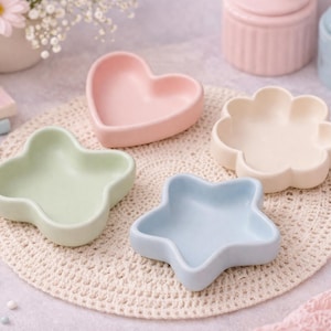 Puede incluir: Conjunto de cuencos de cerámica en colores pastel, con formas de corazón, flor, estrella y trébol de cuatro hojas. Los cuencos están colocados sobre un tapete de ganchillo color crema. Otros elementos incluyen pequeños tarros, libros y flores.