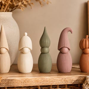 Puede incluir: Un conjunto de cinco figuras decorativas con texturas acanaladas en varias formas y colores, incluyendo un gnomo beige con sombrero cónico, uno blanco con sombrero redondo, una forma de gota verde salvia, un gnomo malva con sombrero curvo y una calabaza.
