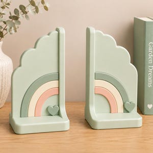 Op de afbeelding: Een paar saliegroene boekensteunen in de vorm van een geschulpte wolk met een gelaagd regenboogontwerp in gedempte groen-, crème- en rozetinten. Elke boekensteun heeft een klein hartje aan de basis van de regenboog voor een stabiele ondersteuning.