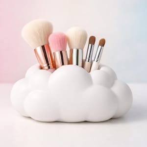 K&ouml;nnte beinhalten: Ein wei&szlig;er, wolkenf&ouml;rmiger Make-up-Pinselhalter, gef&uuml;llt mit verschiedenen Make-up-Pinseln. Die Pinsel haben wei&szlig;e, rosa und braune Borsten mit metallischen Griffen. Der Hintergrund ist ein Farbverlauf aus Rosa und Blau.