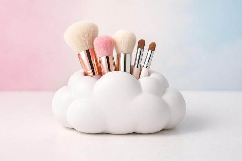 K&ouml;nnte beinhalten: Ein wei&szlig;er, wolkenf&ouml;rmiger Make-up-Pinselhalter, gef&uuml;llt mit verschiedenen Make-up-Pinseln. Die Pinsel haben wei&szlig;e, rosa und braune Borsten mit metallischen Griffen. Der Hintergrund ist ein Farbverlauf aus Rosa und Blau.