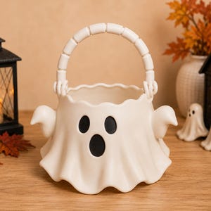 May include: A white ghost-shaped bucket with a scalloped top edge, two small arms extending from the sides, and a black printed face featuring two oval eyes and an oval mouth. The bucket includes a white segmented handle attached to the top rim.