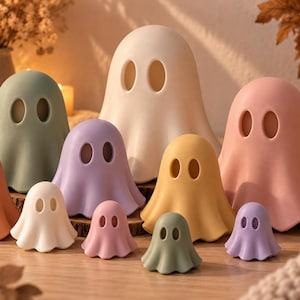 May include: A collection of ten ghost-shaped figurines in various sizes and colours, including white, sage green, terracotta, lavender, yellow, and pink. Each ghost features two oval-shaped eye cutouts and a smooth, rounded, draped silhouette.
