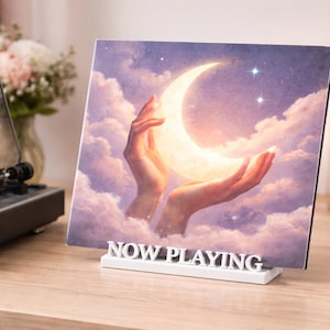 Könnte beinhalten: Ein Vinyl-Schallplattencover mit einem himmlischen Design von Händen, die einen leuchtenden Halbmond vor einem bewölkten Himmel halten. Das Albumcover wird in einem weißen "NOW PLAYING"-Ständer neben einem Plattenspieler und einer brennenden Kerze präsentiert.