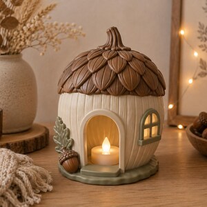 May include: A decorative acorn-shaped house lantern featuring a textured cream-coloured base, a brown cap with leaf detailing, and a small arched doorway. The interior contains a small, glowing white tea light candle, and the exterior is accented with a green leaf and a small acorn near the entrance.