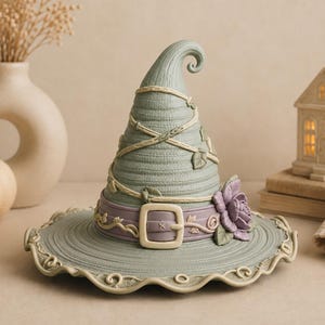 Peut inclure: Une figurine de chapeau de sorcière vert sauge présentant une surface texturée avec des détails de vignes enroulées, une bande violette avec une boucle centrale et une rose violette sculptée. Le bord est ondulé avec des volutes décoratives.