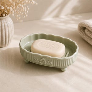 May include: A light green, oval-shaped soap dish with a decorative floral design and a bar of soap. The soap dish has a scalloped edge and is supported by small feet. The soap has the text "SAVON EXTRA DOUX PARIS".