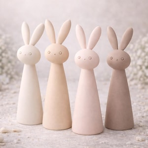 Puede incluir: Cuatro figuras de conejos en colores pastel. Cada una tiene un cuerpo cónico y una cabeza redondeada con ojos pequeños y circulares y orejas largas. Los colores van del blanco roto al marrón claro. El fondo presenta flores blancas y pequeñas piedras redondas.