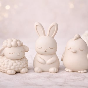 May include: Three cream-colored figurines: a sheep, a bunny, and a chick. The sheep has a textured wool coat, the bunny has long ears, and the chick has small wings. All three have closed eyes, suggesting a peaceful theme.