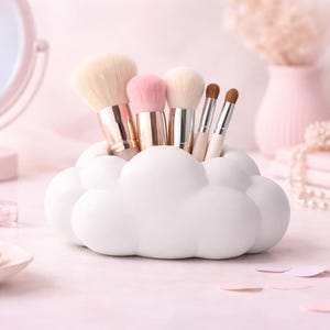 K&ouml;nnte beinhalten: Ein wei&szlig;er, wolkenf&ouml;rmiger Make-up-Pinselhalter, gef&uuml;llt mit verschiedenen Make-up-Pinseln. Die Pinsel haben goldene und silberne Griffe und weiche Borsten. Die Szene spielt auf einer rosa Oberfl&auml;che mit anderem Sch&ouml;nheitszubeh&ouml;r.