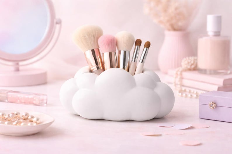 K&ouml;nnte beinhalten: Ein wei&szlig;er, wolkenf&ouml;rmiger Make-up-Pinselhalter, gef&uuml;llt mit verschiedenen Make-up-Pinseln. Die Pinsel haben goldene und silberne Griffe und weiche Borsten. Die Szene spielt auf einer rosa Oberfl&auml;che mit anderem Sch&ouml;nheitszubeh&ouml;r.