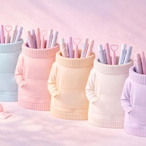 Könnte beinhalten: Fünf pastellfarbene Stifthalter in Form von Pullovern, jeweils gefüllt mit Stiften und herzförmigen Aufsätzen. Die Halter sind in Blau-, Rosa-, Gelb-, Weiß- und Lilatönen gehalten. Eine rosa Schleife liegt im Vordergrund und deutet auf ein Thema von Schreibwaren oder Büroorganisation hin.