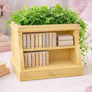 May include: A light yellow wooden bookshelf with two shelves and a drawer, filled with small, off-white books. The top of the bookshelf is filled with green foliage. A teacup, books, and glasses are in the background.