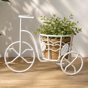 Può includere: Un supporto per piante in metallo bianco a forma di bicicletta con un cestino decorato da dettagli a forma di foglia. All'interno del cestino si trova un vaso rotondo avvolto in corda di juta naturale, contenente una piccola pianta verde.