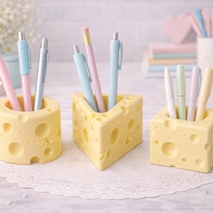 May include: Three yellow cheese-shaped desk organizers with various pens and pencils inside. The organizers have round holes and are arranged on a white lace doily. Pens are pastel pink, blue, and silver.