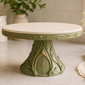 Puede incluir: Un soporte para pasteles de color verde salvia con una parte superior circular blanca y plana. La base presenta un diseño de arco esculpido con detalles pintados en oro, relieves intrincados de enredaderas y una pequeña estrella.