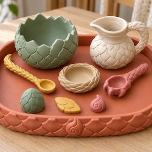 May include: A terracotta-colored tray with a scale pattern holds a sage green egg-shaped bowl, a cream-colored pitcher with a textured handle, a small woven-style bowl, two spoons, a green egg, a yellow leaf, and a small pink swirl.