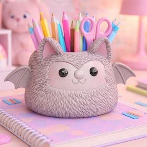 May include: A gray bat-shaped pencil holder with pink ears and wings, filled with colorful pencils and scissors. It sits on a spiral-bound notebook with a pastel cover, alongside other stationery items.