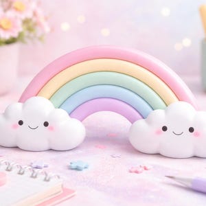 Könnte beinhalten: Ein pastellfarbener Regenbogen mit lächelnden Wolkenakzenten. Der Regenbogen hat Bänder in Rosa, Gelb, Grün, Blau und Lila. Zwei weiße Wolken mit lächelnden Gesichtern sitzen darunter. Die Szene spielt auf einer rosa Oberfläche mit einem Notizbuch und einem Bleistift.