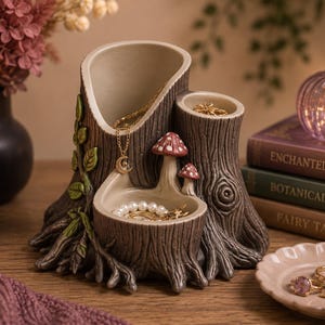May include: A gray tree stump-shaped jewelry organizer featuring two deep cylindrical compartments and a shallow front basin. The exterior is textured like bark with green vine accents and two small red-capped mushrooms on the front ledge.