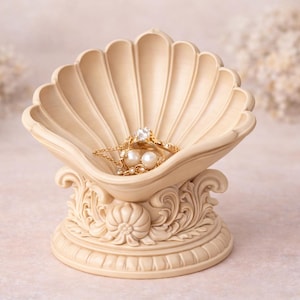 May include: A beige seashell-shaped jewelry dish with a detailed base, holding a gold ring and necklace with pearls. The dish has a textured surface and is set against a soft, blurred background.