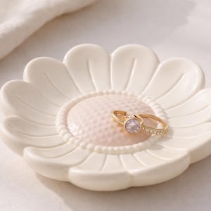 May include: A cream-coloured, daisy-shaped ring dish with a textured pink centre. Two gold rings with gemstones rest on the dish. The dish is on a white surface, with a white cloth in the background.