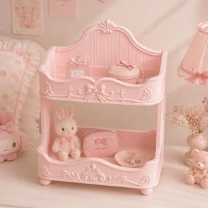 Puede incluir: Estantería decorativa rosa de dos niveles con detalles ornamentados, que contiene un pequeño conejito de peluche, un bolso rosa y un plato con perlas. La estantería superior muestra un frasco de perfume y una caja redonda. La estantería está sobre una superficie blanca.