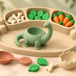 May include: A beige divided tray featuring a central green dinosaur-shaped bowl, surrounded by small beige bone-shaped pieces, green leaf-shaped pieces, and orange carrot-shaped pieces. Includes a terracotta leaf-shaped spoon and a beige eggshell bowl.