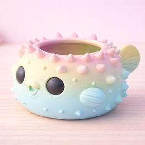 May include: A pastel-colored, round ceramic planter shaped like a pufferfish. The planter features a gradient of colors, including pink, yellow, and blue, with black eyes and a fin. The top edge has pink spikes.