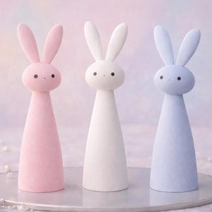 May include: Three pastel-colored bunny figurines. The bunnies are pink, white, and blue, each with a conical body and long ears. They have small black dot eyes and are displayed on a clear, round surface.