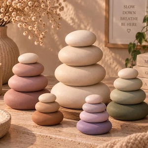 Könnte beinhalten: Fünf Stapel aus glatten, abgerundeten Steinen in verschiedenen Größen und Farben, darunter Creme, Beige, Mauve, Braun, Lavendel und Salbeigrün. Jeder Stapel ist vertikal angeordnet, mit dem kleinsten Stein oben und dem größten Stein an der Basis.