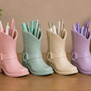 Peut inclure: Quatre porte-stylos en forme de bottes de cow-boy en rose pastel, vert menthe, crème et lavande. Chaque botte présente une sangle décorative avec une boucle et contient une collection de stylos et de crayons aux couleurs assorties.