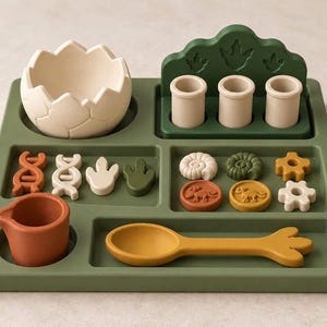 May include: A green compartmentalized tray containing a cream-colored cracked egg, three small cylinders, a yellow spoon, and various small silicone shapes including DNA strands, dinosaur footprints, ammonites, and dinosaur-themed coins.