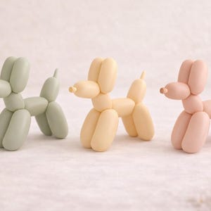 Può includere: Tre sculture di cani a palloncino in colori pastello: verde salvia, giallo pallido e rosa tenue. Ogni cane è progettato per assomigliare a un animale fatto con i palloncini, con forme arrotondate e una piccola coda. Le sculture sono esposte su una superficie bianca.
