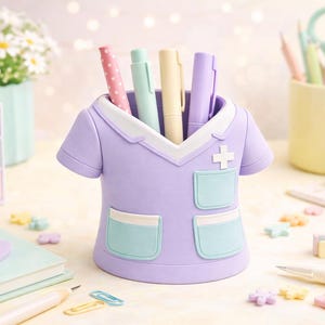 Peut inclure: Un organiseur de bureau en forme de blouse d'infirmi&egrave;re lilas contient des stylos et des marqueurs. Il pr&eacute;sente un col blanc, une croix et deux poches avec une bordure bleu clair. D'autres articles de papeterie et un petit arrangement floral sont en arri&egrave;re-plan.