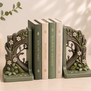 May include: A pair of dark green bookends featuring a sculpted tree trunk design with white flowers and green leaves, holding four books titled Into the Forest, Wildwood, Botanical Journal, and Tales from the Grove with gold leaf accents.