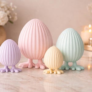 Puede incluir: Colección de figuras decorativas con forma de huevo en colores pastel: lavanda, rosa, amarillo y verde menta. Cada huevo tiene una textura acanalada y pequeñas bases en forma de pie. Las figuras están dispuestas sobre una superficie de mármol.