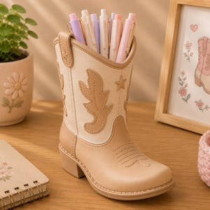 May include: A beige and cream-colored cowboy boot-shaped desk organizer featuring stitched leaf and star patterns on the shaft. The boot is filled with several pastel-colored pens and serves as a functional container for office supplies.