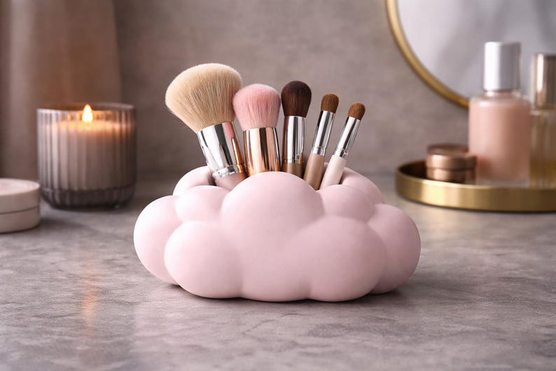 K&ouml;nnte beinhalten: Ein rosa wolkenf&ouml;rmiger Make-up-Pinselhalter mit verschiedenen Pinseln. Der Halter besteht aus Keramik und steht auf einer grauen Oberfl&auml;che. Im Hintergrund befinden sich eine Kerze, ein Spiegel und Sch&ouml;nheitsprodukte.